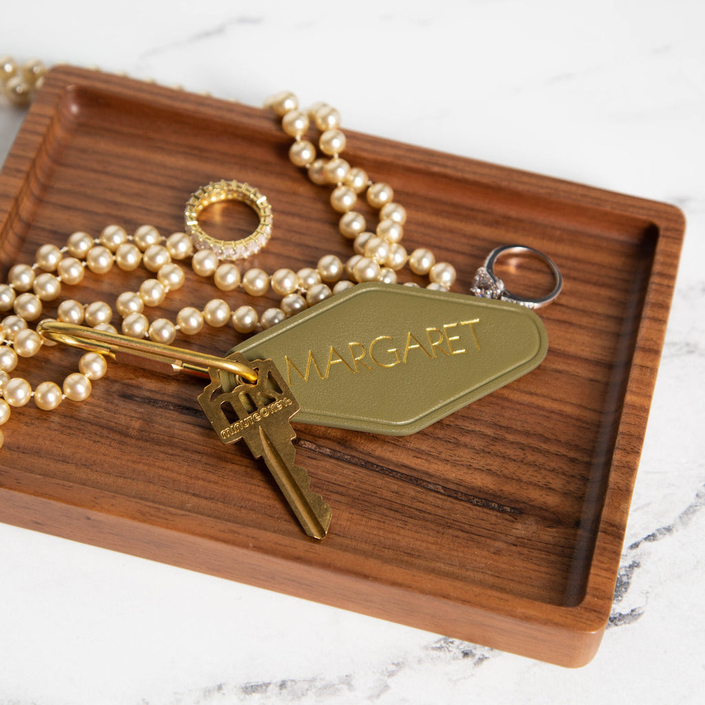 May include: Olive green leather key chain with gold lettering that says &#39;MARGARET&#39;.  The keychain is attached to a gold-toned key.  A gold ring and a pearl necklace are also in the image, resting on a dark brown wooden tray.