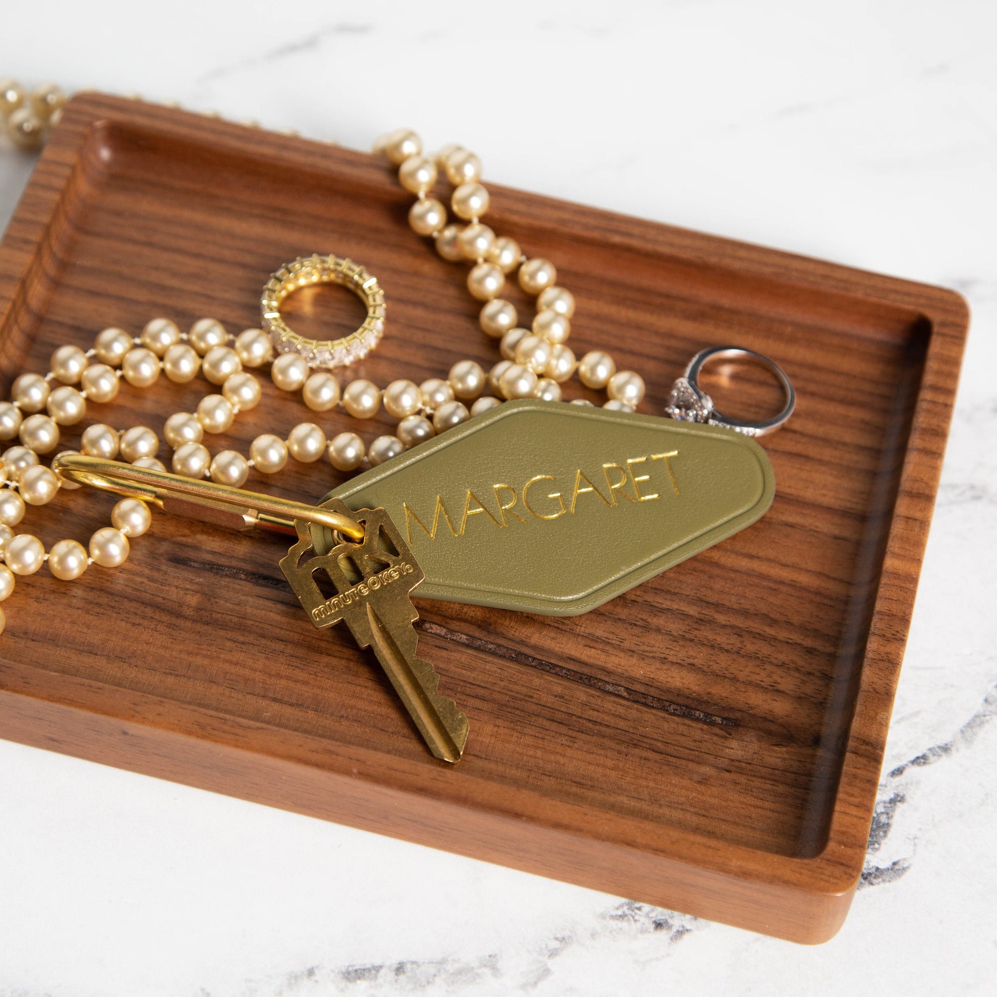 May include: Olive green leather key chain with gold lettering that says &#39;MARGARET&#39;.  The keychain is attached to a gold-toned key.  A gold ring and a pearl necklace are also in the image, resting on a dark brown wooden tray.