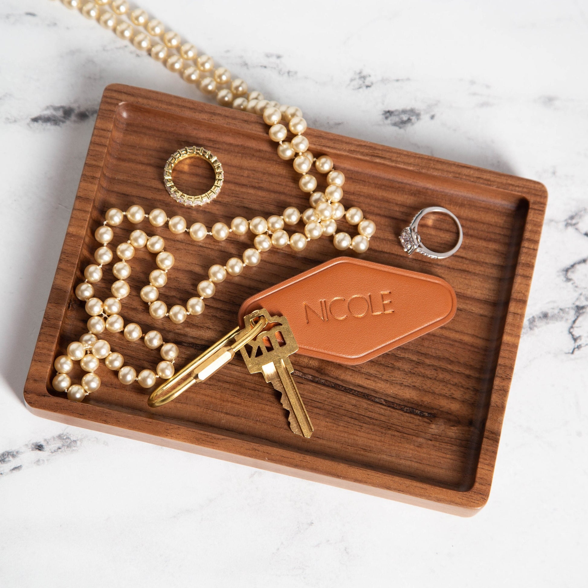 May include: Tan leather key holder with the name &#39;Nicole&#39; embossed on it, attached to a gold-toned key.  A pearl necklace, gold ring, and silver ring are also displayed on a dark brown wooden tray.