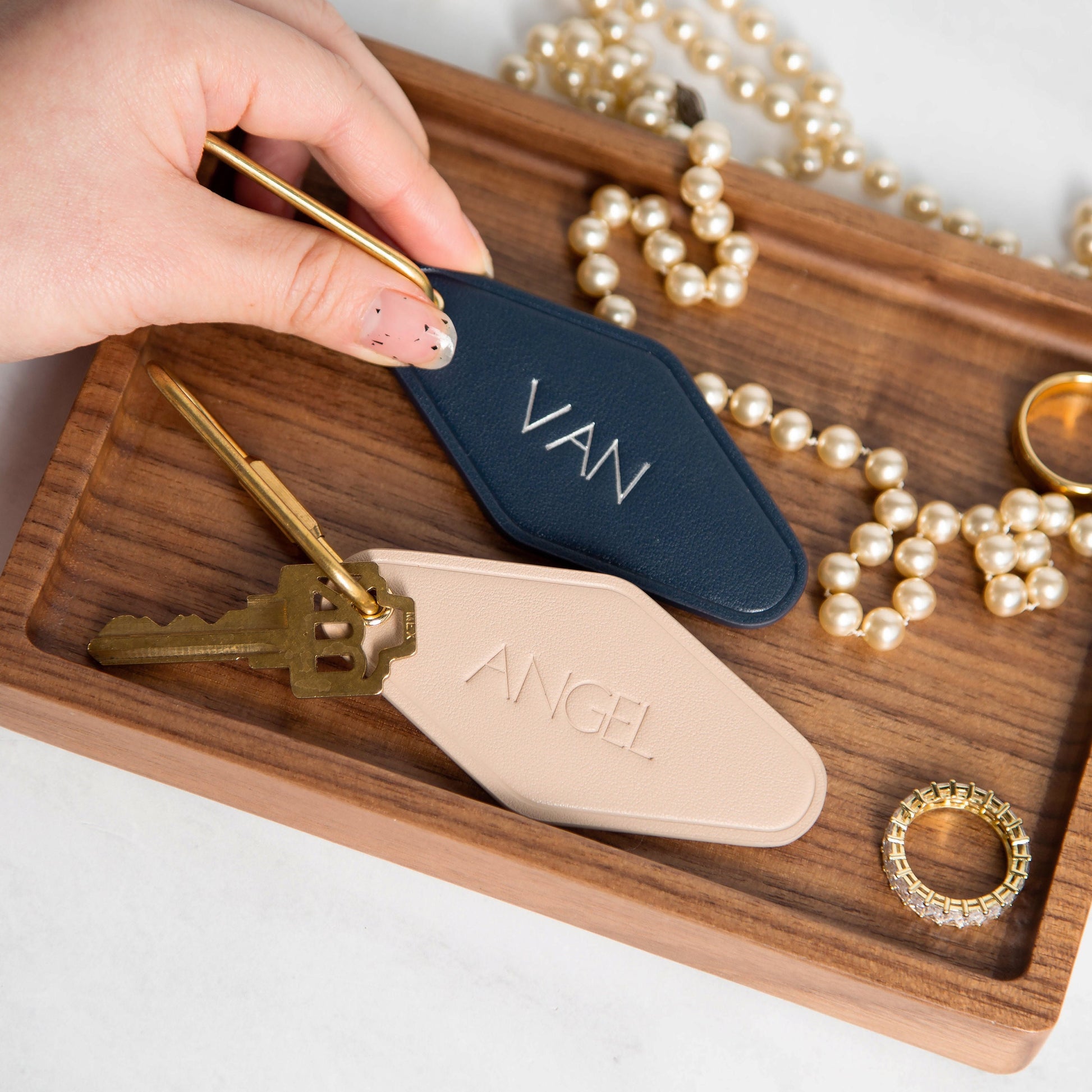 May include: Two personalized leather keychains, one navy blue and one beige, in a luggage tag shape.  The navy blue keychain is engraved with &#39;VAN&#39; and the beige keychain is engraved with &#39;ANGEL&#39;. Both keychains are attached to brass-colored keys.  The keychains are displayed on a wooden tray with pearl necklaces and rings.